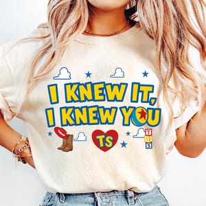 May include: A cream-colored t-shirt featuring the text I KNEW IT, I KNEW YOU in yellow with blue outlines. The design includes a red cowboy hat, brown cowboy boots, a red heart with TS, a yellow ball with a red star, and alphabet blocks.