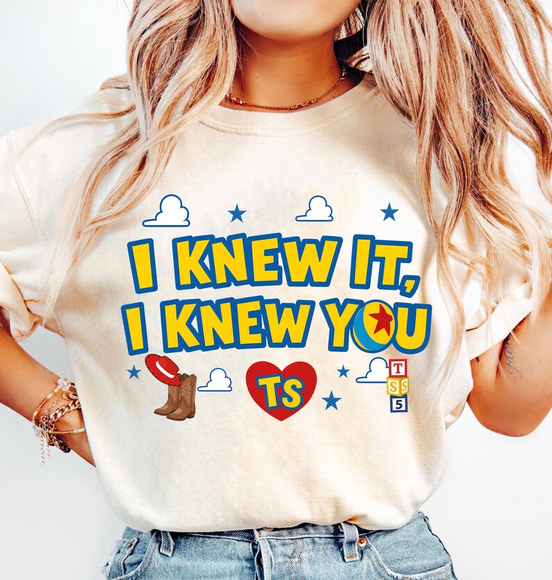 May include: A cream-colored t-shirt featuring the text I KNEW IT, I KNEW YOU in yellow with blue outlines. The design includes a red cowboy hat, brown cowboy boots, a red heart with TS, a yellow ball with a red star, and alphabet blocks.