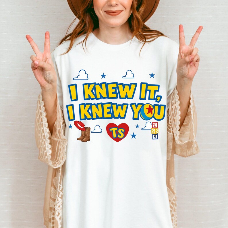 May include: A white t-shirt featuring the blue and yellow text I KNEW IT, I KNEW YOU. The design includes a red cowboy hat, brown boots, a red heart with the letters TS, a colorful ball, blue stars, white clouds, and a stack of lettered blocks.