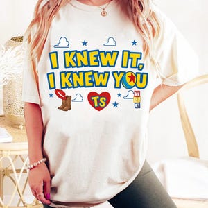 May include: A cream-colored short-sleeve t-shirt featuring the blue and yellow text I KNEW IT, I KNEW YOU with a red heart containing the letters TS, a red cowboy hat, brown boots, blue stars, white cloud outlines, and a stack of alphabet blocks.