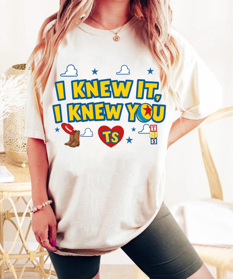May include: A cream-colored short-sleeve t-shirt featuring the blue and yellow text I KNEW IT, I KNEW YOU with a red heart containing the letters TS, a red cowboy hat, brown boots, blue stars, white cloud outlines, and a stack of alphabet blocks.