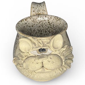 Könnte beinhalten: Eine beige Keramiktasse mit einem Katzen-Gesicht-Design. Die Tasse hat einen gesprenkelten Innenraum und Henkel. Das Katzengesicht hat große, runde Wangen, eine kleine Nase und detaillierte Augen. Die Tasse ist etwa 10 cm hoch.
