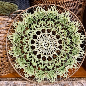 May include: A circular dream catcher with a detailed, openwork pattern. The centre is a light cream colour, fading to green shades towards the edges. The dream catcher is approximately 30 cm in diameter and is made with yarn and a natural-coloured frame.
