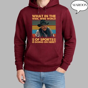 May include: Maroon hoodie with a graphic featuring a man in a cowboy hat and the text "WHAT IN THE WIDE, WIDE WORLD OF SPORTS IS GOING ON HERE?". The word "MAROON" is in a speech bubble.