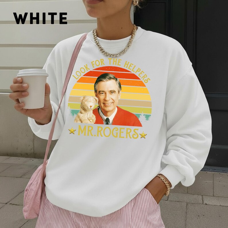May include: White sweatshirt featuring a graphic of Fred Rogers with a puppet and the text "Look for the Helpers" and "Mr. Rogers" in yellow. The word "WHITE" is printed in black at the top left.
