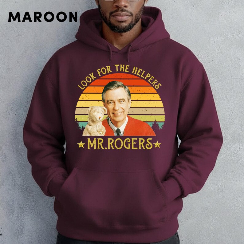May include: Maroon hoodie featuring a graphic of Mr. Rogers with the text "LOOK FOR THE HELPERS" and "MR. ROGERS" in a vintage style. The design includes a sunset and a stuffed animal. The hoodie has a drawstring hood and a front pocket.