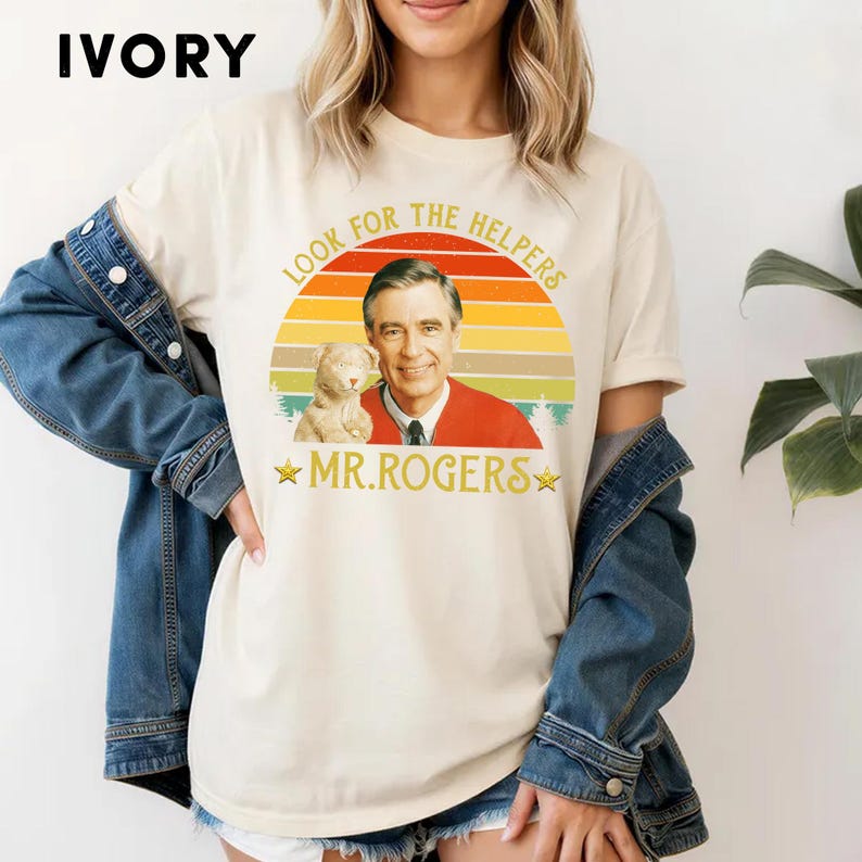 May include: Ivory-colored t-shirt featuring a graphic of Mr. Rogers with a cat and the text "Look for the Helpers" and "Mr. Rogers" in a retro design. The shirt has short sleeves and a crew neck.