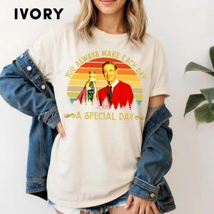 May include: Ivory-colored t-shirt featuring a graphic of a man in a red jacket and a figure in a crown, set against a sunset design. The text reads "YOU ALWAYS MAKE EACH DAY A SPECIAL DAY."