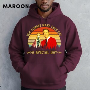 May include: Maroon hoodie featuring a graphic of a man in a red sweater and a puppet, with the text "YOU ALWAYS MAKE EACH DAY A SPECIAL DAY" in gold lettering. The design is set against a sunset background.