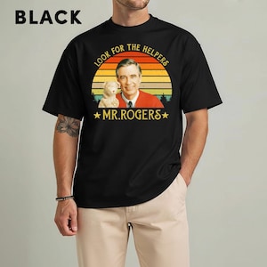 May include: Black t-shirt featuring a graphic of Mr. Rogers with a puppet and the text "LOOK FOR THE HELPERS" and "MR. ROGERS" in a retro design. The shirt is a crew neck and has short sleeves.