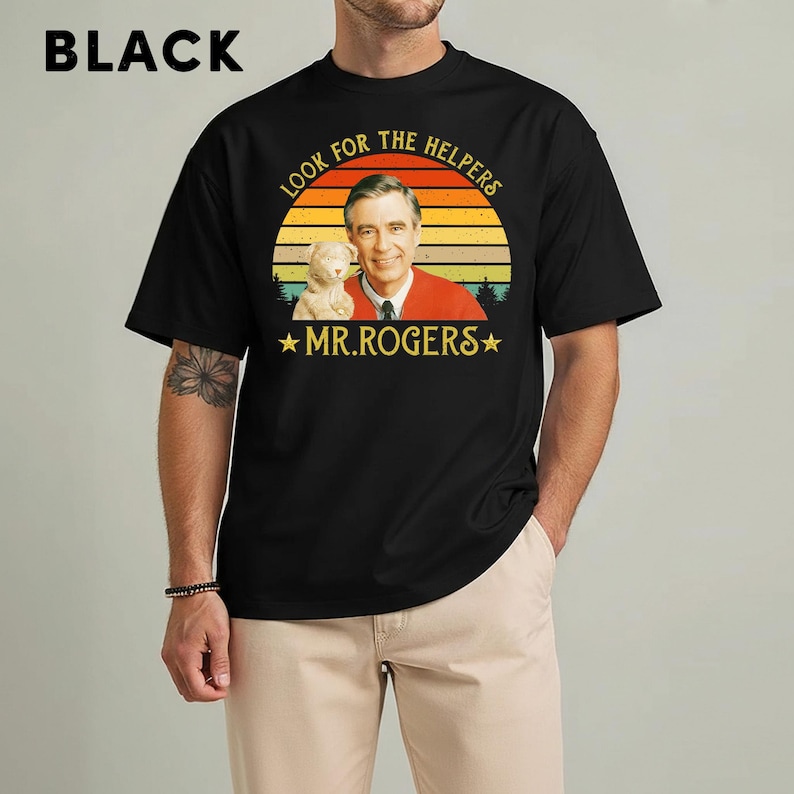 May include: Black t-shirt featuring a graphic of Mr. Rogers with a puppet and the text "LOOK FOR THE HELPERS" and "MR. ROGERS" in a retro design. The shirt is a crew neck and has short sleeves.