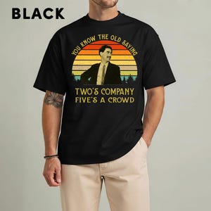 May include: Black t-shirt with a graphic featuring a man in a suit against a sunset backdrop. The text reads "YOU KNOW THE OLD SAYING" above and "TWO'S COMPANY FIVE'S A CROWD" below. The word "BLACK" is at the top.