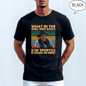 May include: Black t-shirt with a graphic featuring a man in a cowboy hat and the text "WHAT IN THE WIDE, WIDE WORLD OF SPORTS IS GOING ON HERE?". The design includes a retro color scheme with blue, green, and orange stripes.