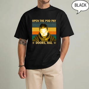 May include: Black t-shirt with a retro design featuring the text "OPEN THE POD PAY DOORS, HAL" and an image of a person. The design includes stripes in green, orange, and yellow. The word "BLACK" is in a speech bubble.