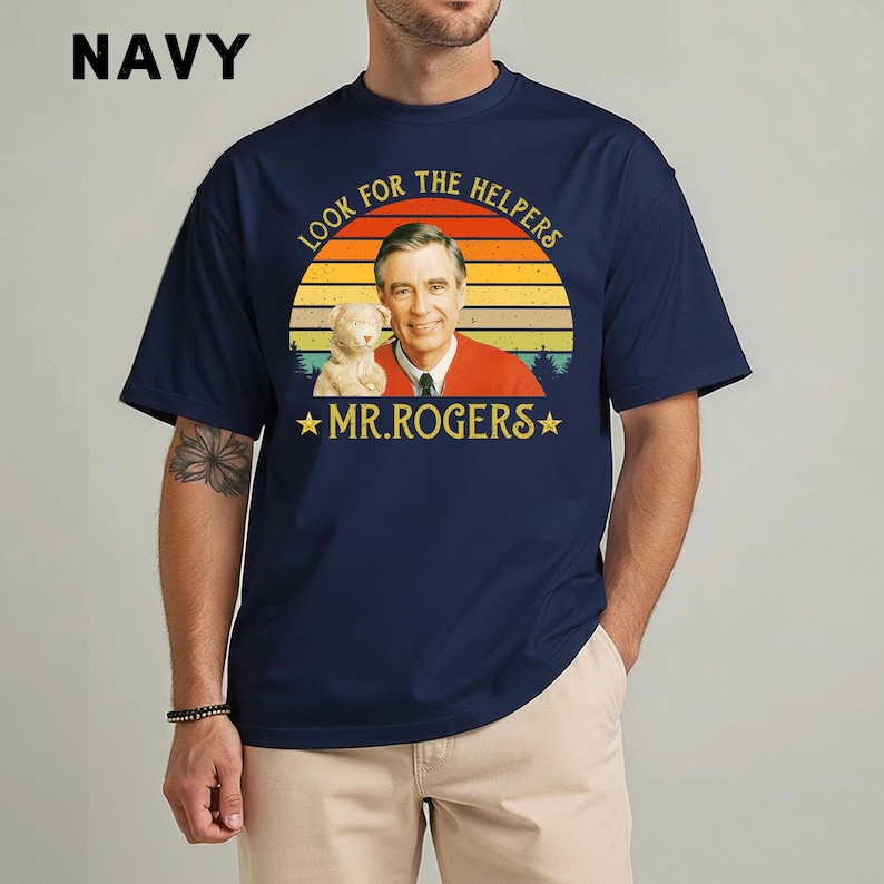 May include: Navy blue t-shirt featuring a graphic of Mr. Rogers with the text "LOOK FOR THE HELPERS" and "MR. ROGERS" in a retro design. The shirt has a crew neck and short sleeves. The word "NAVY" is printed in white in the upper left corner.