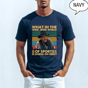 May include: Navy blue t-shirt featuring a graphic with the text "WHAT IN THE WIDE, WIDE WORLD OF SPORTS IS GOING ON HERE?". The graphic includes an image of a man wearing a cowboy hat. The word "NAVY" is in a speech bubble.