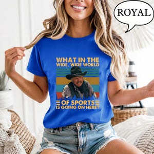 May include: Royal blue t-shirt with a graphic featuring a cowboy and the text "WHAT IN THE WIDE, WIDE WORLD OF SPORTS IS GOING ON HERE?". The design includes a cowboy hat and a retro color scheme.