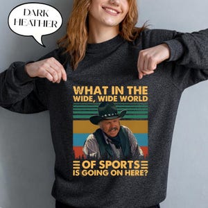 May include: Dark heather gray sweatshirt with a graphic featuring a cowboy and the text "WHAT IN THE WIDE, WIDE WORLD OF SPORTS IS GOING ON HERE?". The sweatshirt has a crew neck and long sleeves.