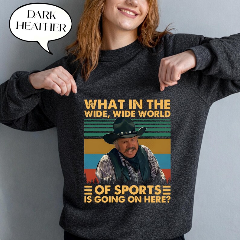May include: Dark heather gray sweatshirt with a graphic featuring a cowboy and the text "WHAT IN THE WIDE, WIDE WORLD OF SPORTS IS GOING ON HERE?". The sweatshirt has a crew neck and long sleeves.