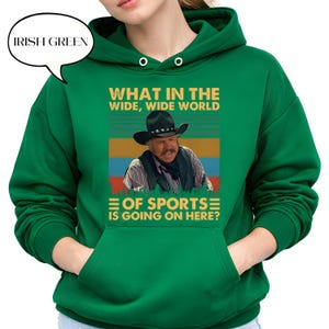 May include: Irish green hoodie with a graphic featuring a man in a cowboy hat and the text "WHAT IN THE WIDE, WIDE WORLD OF SPORTS IS GOING ON HERE?". The hoodie has a front pocket and a drawstring hood.