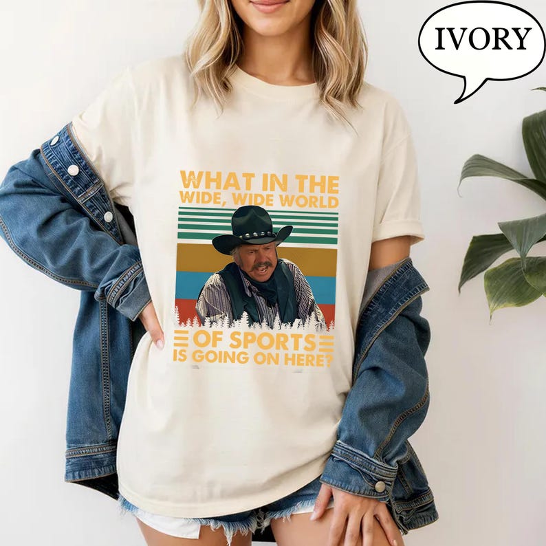 May include: Ivory-colored t-shirt featuring a graphic of a man in a cowboy hat with the text "WHAT IN THE WIDE, WIDE WORLD OF SPORTS IS GOING ON HERE". The design includes a striped background in green, brown, and blue.