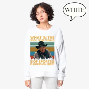 May include: White sweatshirt featuring a graphic of a man in a cowboy hat with the text "WHAT IN THE WIDE, WIDE WORLD OF SPORTS IS GOING ON HERE?". The design includes a colorful striped background and a speech bubble that says "WHITE".