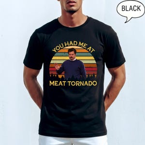 May include: Black t-shirt featuring a retro sunset design with the text "YOU HAD ME AT MEAT TORNADO." The graphic includes an image of a man in a blue sweater. The word "BLACK" is in a speech bubble.