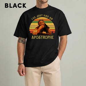 May include: Black t-shirt with a graphic of a man eating, set against a sunset design. The text reads "I'VE JUST HAD AN APOSTROPHE" in yellow. The word "BLACK" is printed in white at the top left.