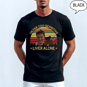 May include: A black short-sleeve t-shirt featuring a graphic print of a person holding a handgun. The design includes a retro-style sunset background with horizontal stripes and the yellow text "I'M FAR TOO SENSITIVE LIVER ALONE" with stars.