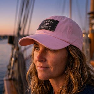 May include: A light pink baseball cap with a curved brim. The cap features a black rectangular patch with the words "OLD SALT NANTUCKET EST. 1859" in white. The cap is made of a soft material.