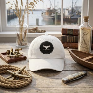 May include: A white baseball cap with a black circular patch featuring a whale tail design. The hat is displayed on a wooden surface with nautical-themed items, including a small boat, rope, and a vintage map in a bottle. The background shows a harbor scene.