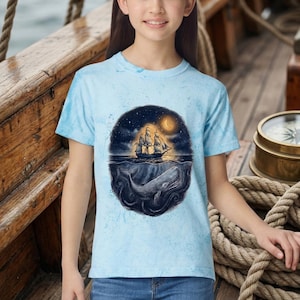 May include: A light blue t-shirt featuring a nautical design. The graphic depicts a sailing ship under a starry night sky with a crescent moon, above a whale in the ocean. The shirt has a tie-dye effect.