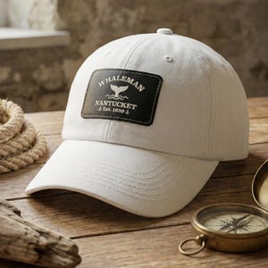 Puede incluir: Gorra de béisbol blanca con visera curva. La gorra presenta un parche negro con el texto "Whaleman Nantucket Est. 1659" y un gráfico de cola de ballena. Un compás sugiere un tema náutico. La gorra es de material resistente.