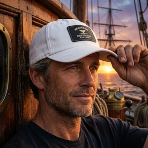 May include: White baseball cap with a black patch that reads "SEAFARER NANTUCKET Est. 1856" and a whale graphic. The cap is worn by a person on a boat, with a sunset in the background.