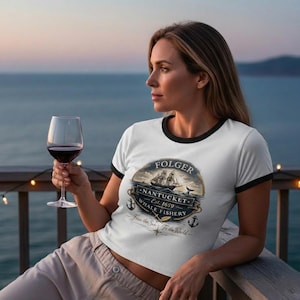 Puede incluir: Camiseta blanca con ribete negro con un diseño náutico. El gráfico incluye un barco, una ballena y el texto "FOLGER NANTUCKET WHALE FISHERY Est. 1659". Una persona lleva la camiseta y sostiene una copa de vino tinto.