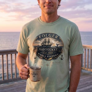 Puede incluir: Camiseta verde salvia con un diseño náutico que presenta un barco, una cola de ballena y el texto "Nantucket Whale Fishery Est. 1659". Una taza marrón de Folger's se sostiene en la mano. La camiseta tiene una estética vintage.