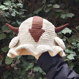 Puede incluir: Un sombrero de pescador de ganchillo color crema con un diseño de flecha marrón en la parte superior, dos cuernos marrones que sobresalen de los lados y pequeños detalles blancos en forma de orejas. Hecho de hilo suave y texturizado con ala ancha.