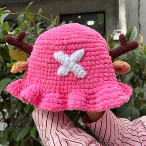 May include: A bright pink crocheted bucket hat featuring a white cross emblem on the front, two brown antlers extending from the sides, and small tan ears positioned below the antlers, all crafted from a soft, textured yarn material.