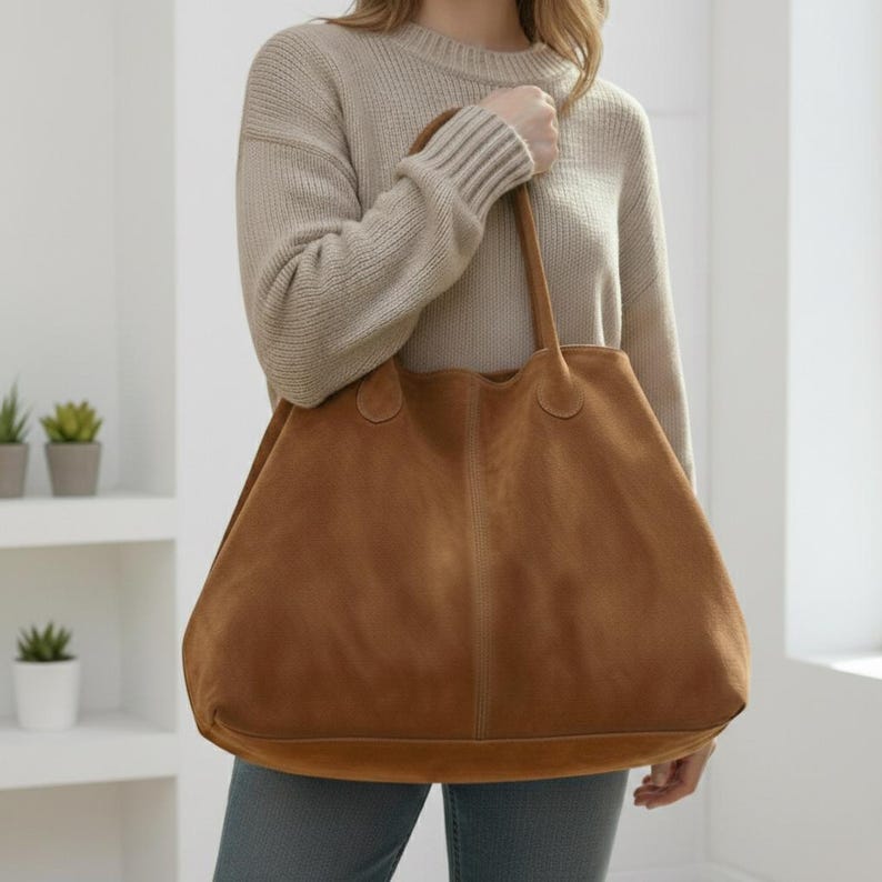 May include: A large, brown suede tote bag with two handles. The bag has a wide, open top and a spacious interior. The bag is being held by a person wearing a beige sweater and blue jeans. The bag is a neutral color and could be used for shopping or everyday use.