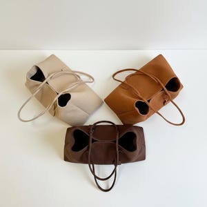May include: Three tote bags in neutral colors: beige, brown, and dark brown. Each bag has a unique design with a bow-like shape and long handles. The bags are made of a soft material and are suitable for everyday use.
