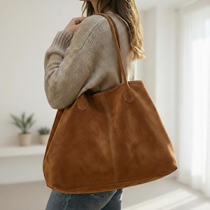 May include: A large, brown suede tote bag with long handles. The bag is a warm, earthy tone and appears soft. The tote is carried over the shoulder. The bag is a simple, classic design.