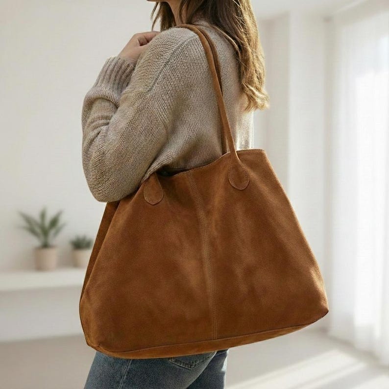 May include: A large, brown suede tote bag with long handles. The bag is a warm, earthy tone and appears soft. The tote is carried over the shoulder. The bag is a simple, classic design.