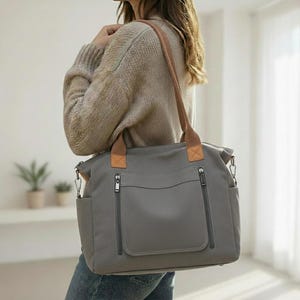 Può includere: Borsa tote in tela grigia con manici e dettagli in pelle marrone. La borsa ha due tasche frontali con cerniera e tasche laterali. La tracolla è in pelle marrone. La borsa viene portata a spalla.