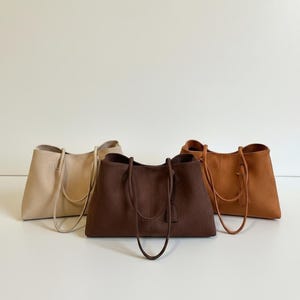 May include: Three tote bags in neutral colors: beige, brown, and tan. Each bag has a rectangular shape with a wide opening and long handles. The bags are made of a soft, textured material, and are suitable for everyday use.