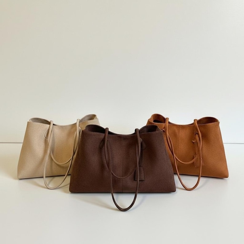May include: Three tote bags in neutral colors: beige, brown, and tan. Each bag has a rectangular shape with a wide opening and long handles. The bags are made of a soft, textured material, and are suitable for everyday use.