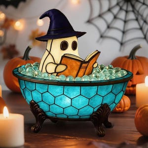 May include: A stained glass lamp shaped like a bathtub with a teal honeycomb pattern and dark metal claw feet. Inside the tub, a cream-colored ghost figure wearing a dark blue pointed hat sits and reads an open brown book.