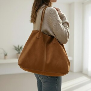 May include: A large, brown suede tote bag with long handles. The bag is carried over the shoulder and has a simple, minimalist design. The bag is a warm, earthy tone and appears to be spacious.