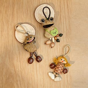 May include: Three handmade wooden ornaments. One is a frog with a green heart-shaped bag, another is a smiling character, and the third is a doll with yellow hair. All have wooden bases and are made with natural materials.