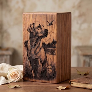 Puede incluir: Una urna rectangular de madera con una ilustraci&oacute;n grabada con l&aacute;ser oscuro de un Labrador sentado en hierba alta cerca de un lago. La escena incluye un p&aacute;jaro volando, monta&ntilde;as lejanas y un bosque. La veta de la madera es visible en la superficie.