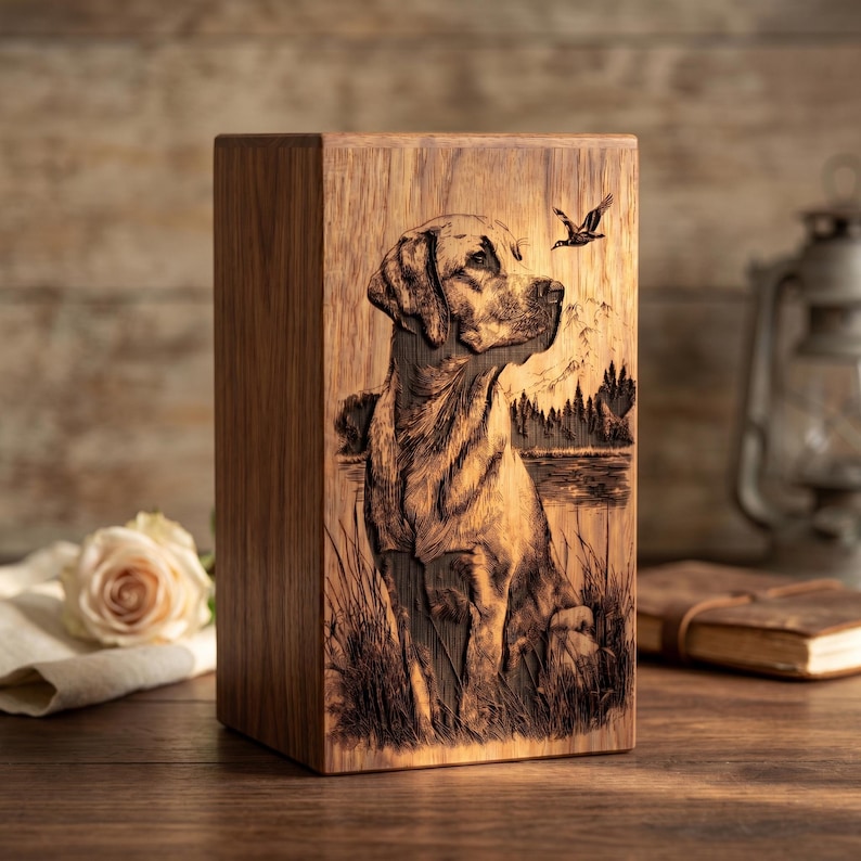 Puede incluir: Una urna rectangular de madera con una imagen grabada con l&aacute;ser de un Labrador sentado en un campo de hierba cerca de un lago. La escena incluye un fondo de bosque, una silueta de monta&ntilde;a y un p&aacute;jaro volando en la esquina superior derecha.